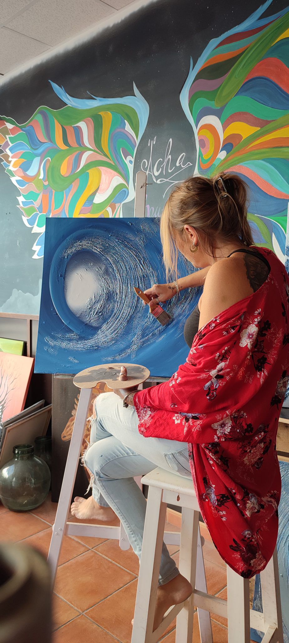 Ächa pintando una ola en su caballete frente a un mural de alas de colores